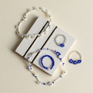 Stilvolle Auswahl an handgefertigtem Perlenschmuck, darunter feine Armbänder mit Smileys und blaue Blumenringe aus Glasperlen auf einer weißen Keramikfliese.