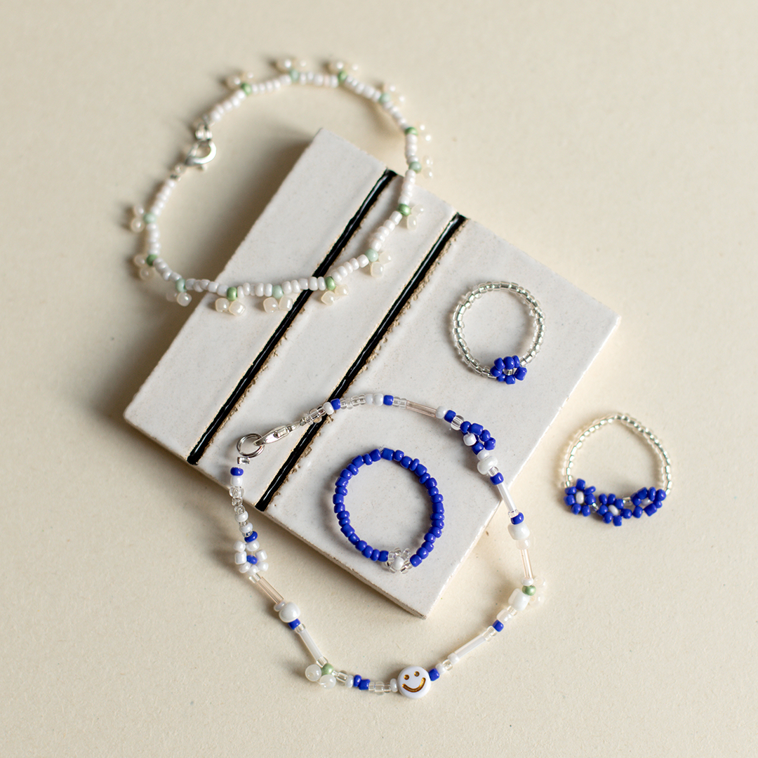 Stilvolle Auswahl an handgefertigtem Perlenschmuck, darunter feine Armbänder mit Smileys und blaue Blumenringe aus Glasperlen auf einer weißen Keramikfliese.