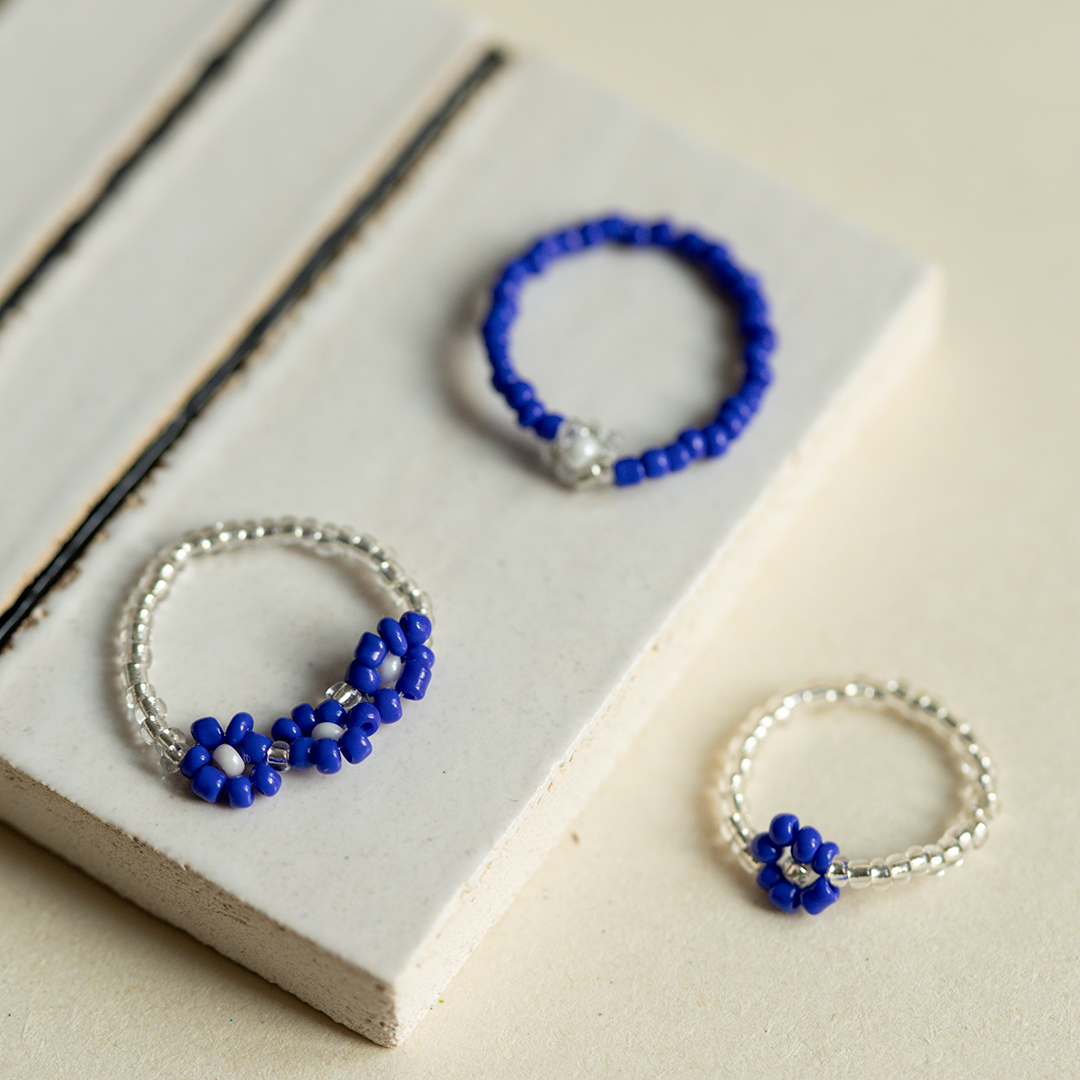 Nahaufnahme von drei feinen, selbstgemachten Ringen aus blauen und transparenten Perlen mit filigranem Vergissmeinnicht-Blumenmuster auf weißem Grund.
