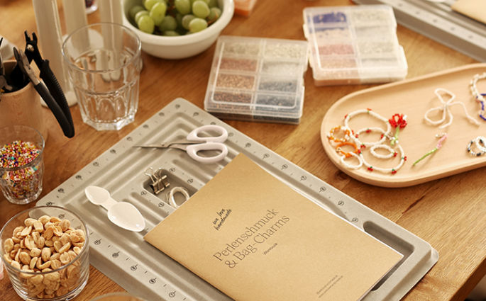 Ein Arbeitstisch im we love handmade Studio für einen Perlenschmuck und Bag Charms Workshop mit Perlenbrettern, Zangen, bunten Perlenboxen, dem Workshop-Workbook und kleinen Snacks wie Nüssen und Weintrauben. | we love handmade