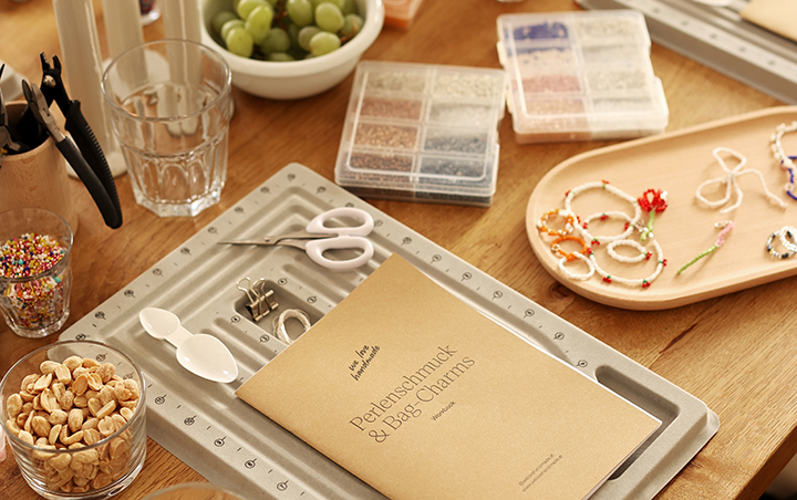 Ein Arbeitstisch im we love handmade Studio für einen Perlenschmuck und Bag Charms Workshop mit Perlenbrettern, Zangen, bunten Perlenboxen, dem Workshop-Workbook und kleinen Snacks wie Nüssen und Weintrauben. | we love handmade