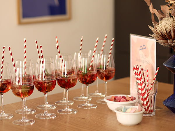 Eine Reihe gefüllter Aperitivo-Gläser mit Beeren und rot-weiß gestreiften Strohhalmen auf einem Holztisch im we love handmade Studio in Wien Neubau. | we love handmade