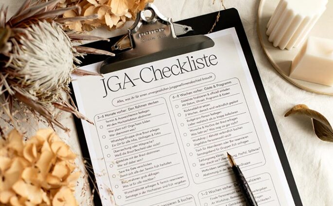 Nahaufnahme im Querformat von der ausgedruckten JGA-Checkliste im Klemmbrett, umrahmt von Trockenblumen und Kerzen.